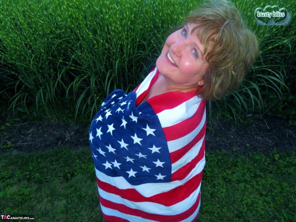 Older plumper Busty Bliss holds an American flag while totally naked in a yard - #15