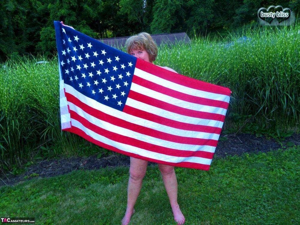 Older plumper Busty Bliss holds an American flag while totally naked in a yard - #12