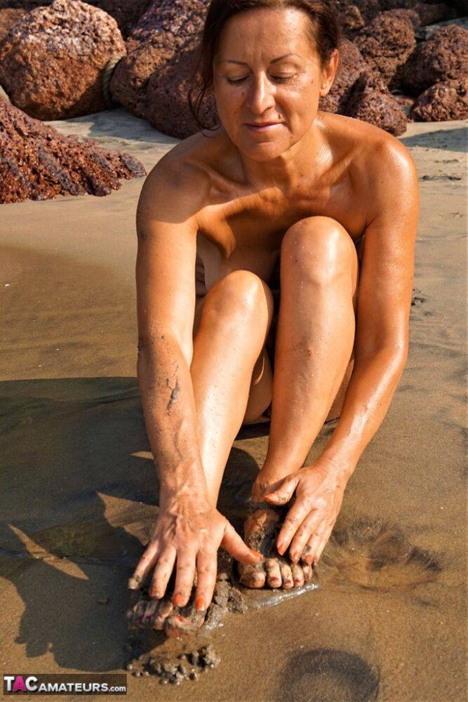 Naked older woman Diana Ananta covers her feet in beach sand at low tide - #7