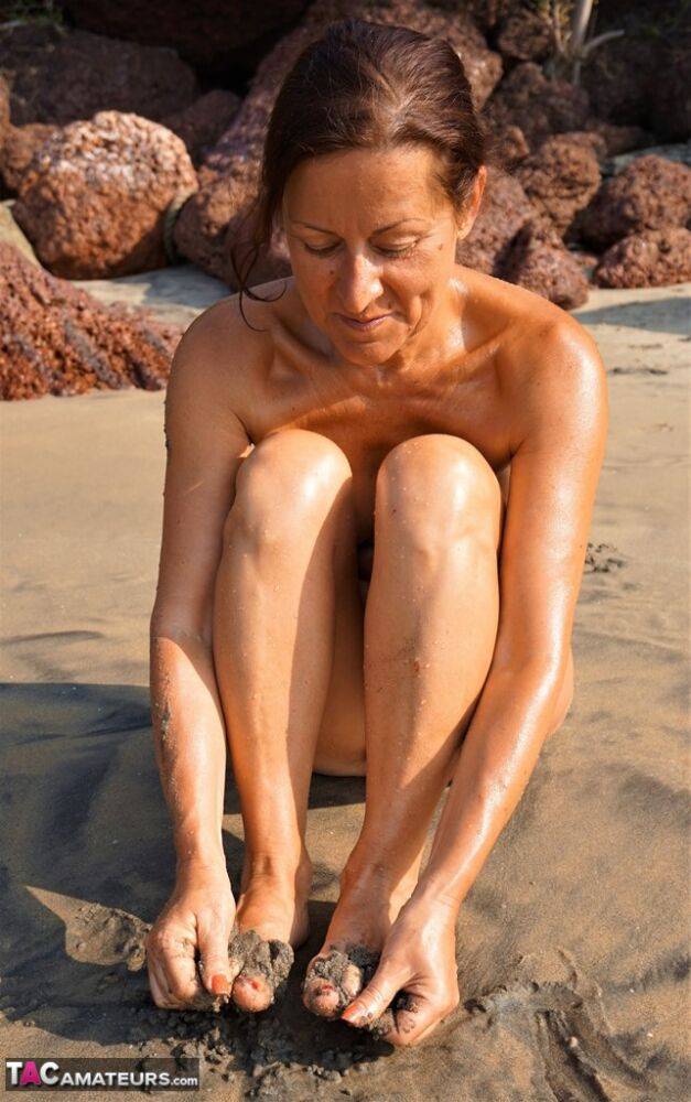 Naked older woman Diana Ananta covers her feet in beach sand at low tide - #2