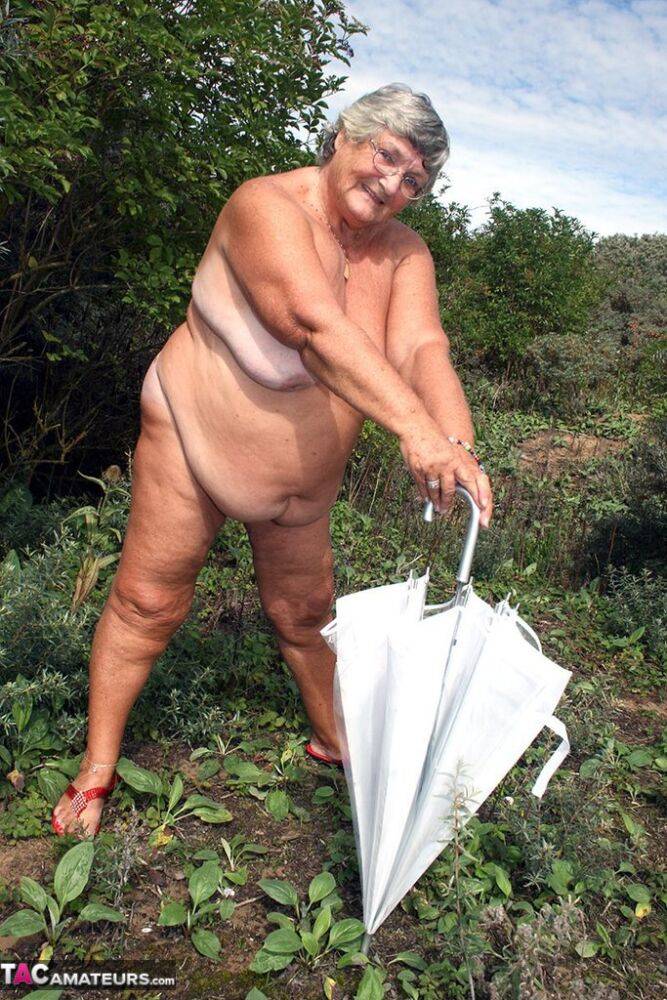 Obese oma Grandma Libby holds an umbrella while posing naked by fir trees - #11