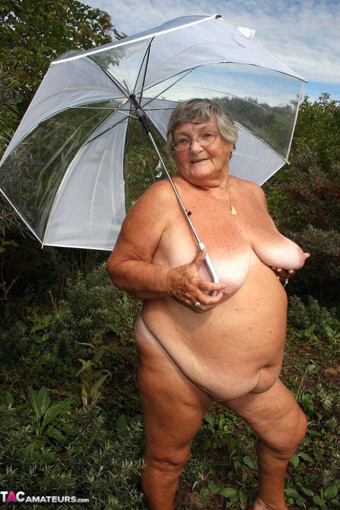 Obese oma Grandma Libby holds an umbrella while posing naked by fir trees - #12