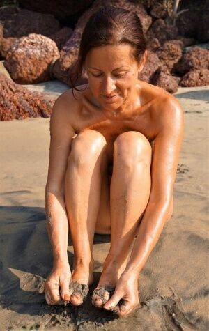 Naked older woman Diana Ananta covers her feet in beach sand at low tide on picsofsex.com